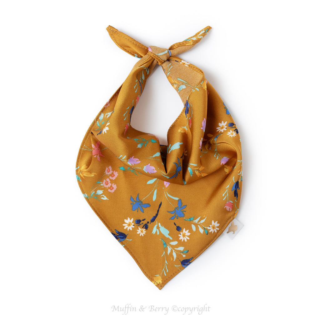 A gold mustard pet bandana with a colorful floral pattern, tied in a loop at the top, shown on a plain white background, by Muffin & Berry