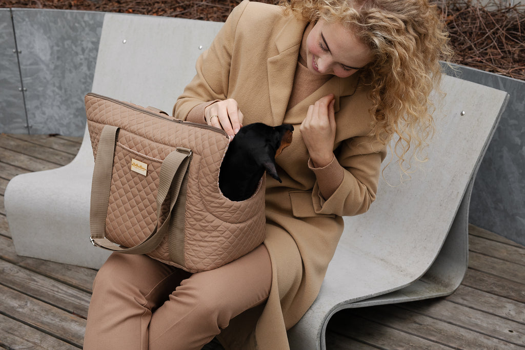 Smiling woman sitting on outdoor bench with AMELIA camel quilted dog carrier bag, Dachshund peeking out, by Muffin & Berry
